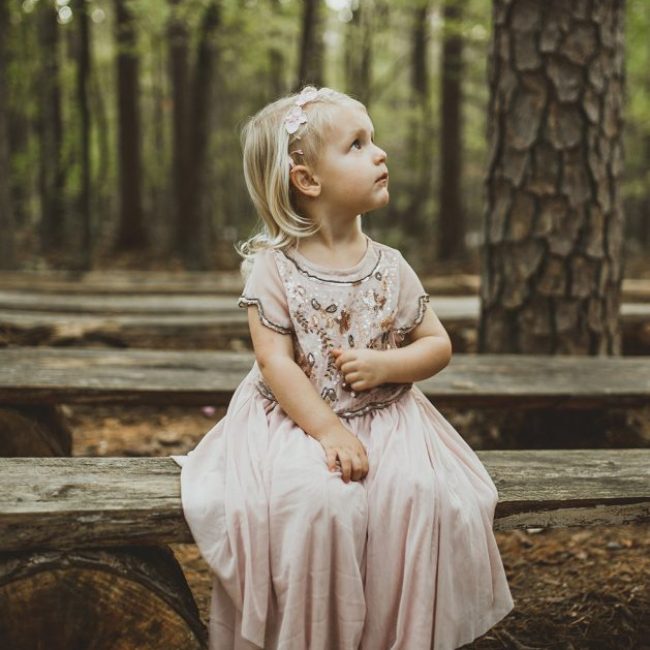 little girl in blush dress sitting on a bench in the woodlands looking up