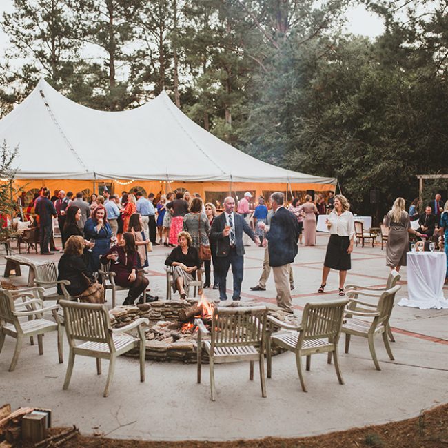 Outdoor wedding reception featuring white tent and courtyard with guests sitting by the fire and socializing